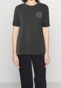 Persona que lleva una camiseta de color gris oscuro con mangas cortas y un pequeño gráfico de sol con el texto "Es un buen día" en la parte superior izquierda del pecho.