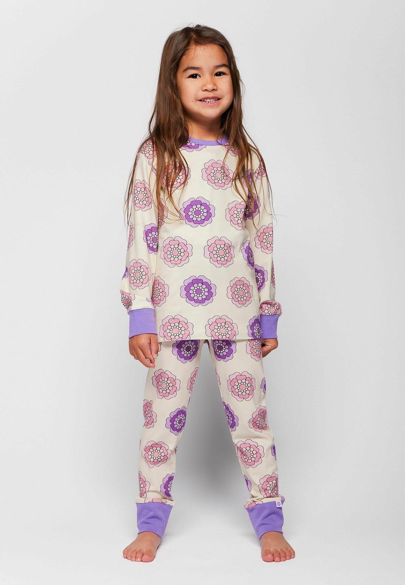 Jeune fille debout pieds nus portant un pyjama à motif floral avec des poignets violets, souriant devant un fond gris clair uni.