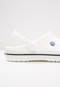 Hvit klogsko med et slankt, lukket design, som har en bakrem, blå logo og en svart stripet såle. Laget av lett materiale.