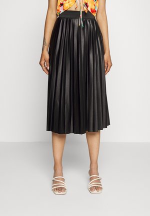 Kobieta ubrana w czarną plisowaną spódnicę midi, kolorowy krótki top i białe sandały na paski, stojąca na tle gładkiej białej ściany.