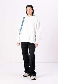 Hvit oversized hettegenser med kengurulomme, kombinert med svarte rette jeans. Akkentuert med en blå stropp på den ene skulderen.