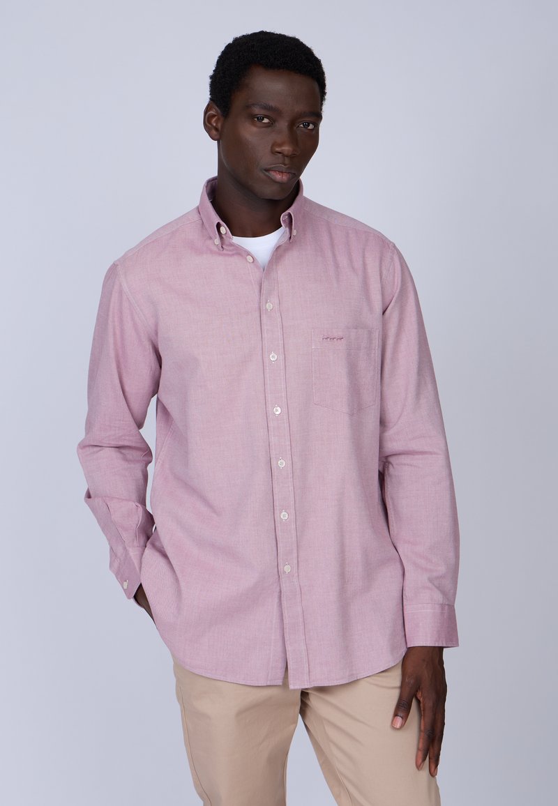 Chemise boutonnière rose clair en mélange de coton avec manches longues, détail de poche et boutons blancs, associée à un pantalon beige.