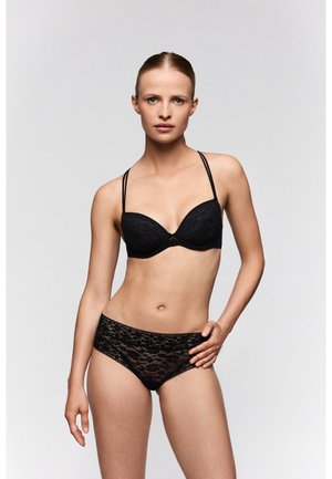 Vrouw die een zwarte kanten beha met dubbele dunne bandjes en bijpassend kanten ondergoed draagt, staand tegen een effen lichte achtergrond.