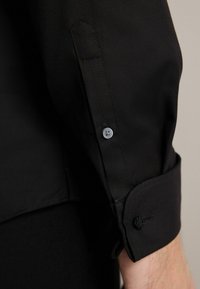 Primăvară închisă, cu mânecă lungă, cu un material texturat, manșetă cu nasture și un detaliu cu nasture. Manșeta are o buclă decorativă pentru fixare sigură.