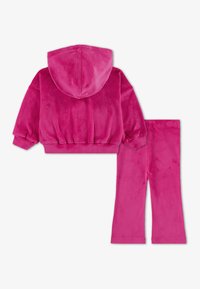 Roze velur trenirka koja se sastoji od dukserice s kapuljačom i rebrastim rubom, te odgovarajućih hlača blago proširenih u zapešćima. Mekana tekstura.