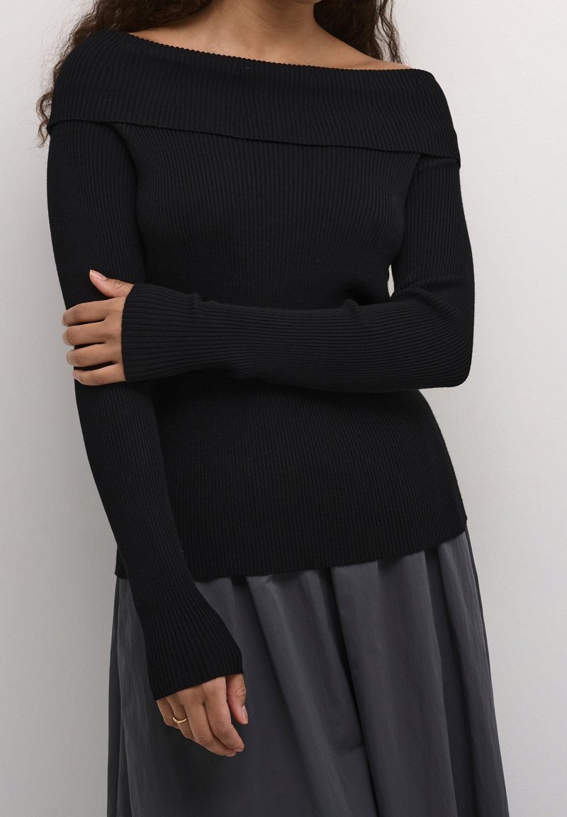 Femme portant un pull noir côtelé à col bateau avec manches longues et une jupe plissée grise, debout devant un fond uni.