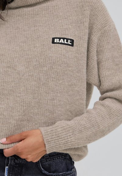Béžový pletený svetr s ribovými manžetami, se černou obdélníkovou nášivkou s nápisem "BALL." Měkká textura a volný střih.