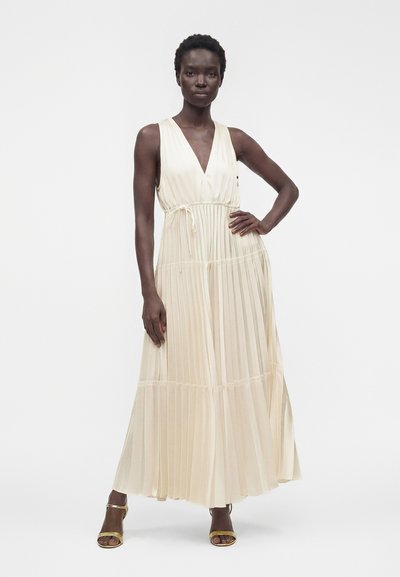 Femme portant une robe longue sans manches plissée couleur crème avec un décolleté en V et une ceinture à la taille, chaussée de sandales à talons hauts dorées.