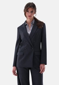 Dunkelgrauer doppelt geknöpfter Blazer mit Revers, zwei vorderen Taschen und glatter Textur, über einem hellgrauen Hemd mit Knopfleiste.