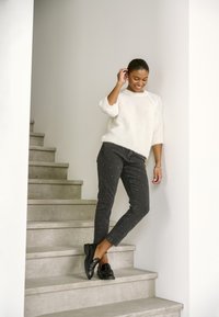 Giovane donna in maglione bianco e jeans neri con borchie, sorridente mentre si trova su scale in cemento all'interno.