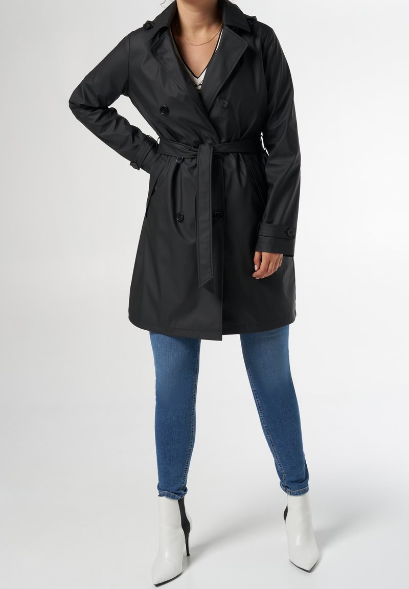 Femme portant un trench-coat noir ceinturé, un jean skinny bleu, des bottes blanches à talons aiguilles, debout devant un fond uni.