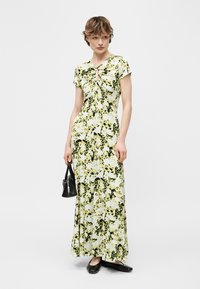 Vrouw draagt een groene en geel bloemige maxi-jurk met korte mouwen, houdt een zwarte handtas vast en draagt zwarte ballerina's tegen een witte achtergrond.