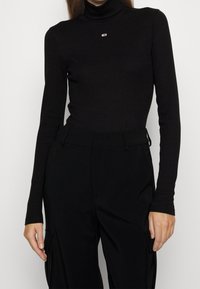 Schwarzes Langarm-Turtleneck-Oberteil aus geripptem Stoff mit kleinem Logo auf der Brust. Kombiniert mit hochgeschnittenen schwarzen Hosen.