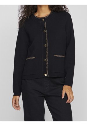 Vrouw draagt een zwart vest met gouden knopen en metalen randen langs de halslijn, knopenrij en zakranden, gecombineerd met een zwarte broek.