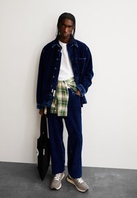Marine corduroy jasje en broek met wit t-shirt, groen geruit overhemd om de taille geknoopt, grijze sneakers en een zwarte tote bag met logo.