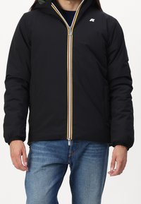 Mand iført en sort hættejakke med frontlynlås, der er trimmet i gule, hvide og røde striber, parret med blå jeans.