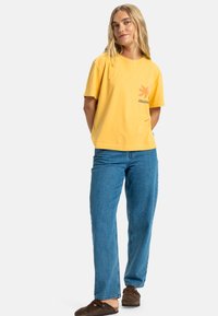 Gul bomull t-skjorte med kort erme, har et grafisk design på venstre bryst. Kombinert med lyseblå denimjeans og brune klomper.