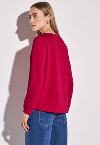 Červená mikina s kulatým výstřihem a dlouhými rukávy, zkombinovaná s světle modrými džíny. Hladká textura, uvolněný střih, minimální detaily.