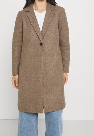 Vrouw die een middellange bruine wollen jas draagt met een enkele zwarte knop, een wit shirt en lichtblauwe spijkerbroek tegen een effen witte achtergrond.