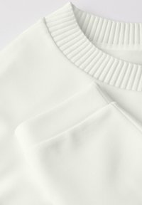 Witte sweatshirt met een geribde ronde hals en een gladde textuur, met opgerolde mouwen netjes op een vlak oppervlak gelegd.