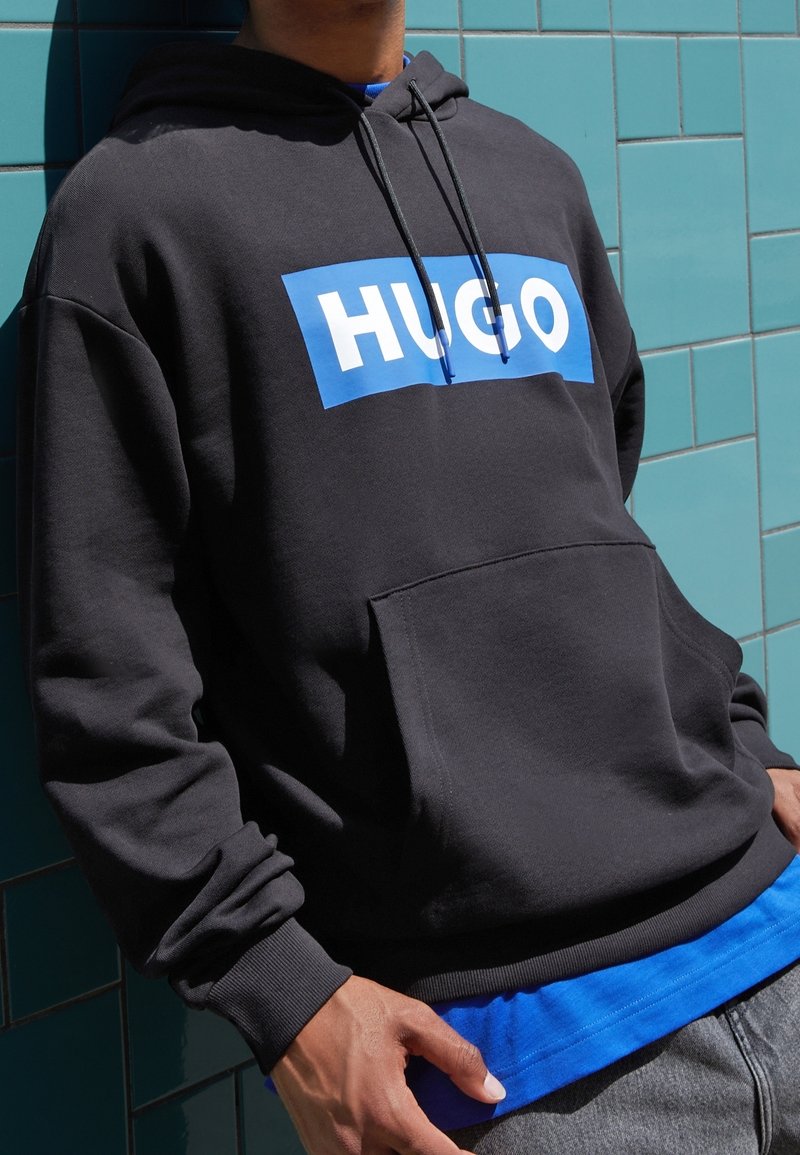 Personne portant un sweat à capuche noir avec un rectangle bleu et un texte blanc "HUGO", debout contre un mur carrelé bleu sarcelle.