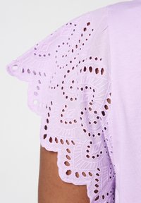 Haut en coton lilas avec des manches en dentelle brodées, ornées de découpes et de bords festonnés. Texture lisse.
