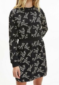 Musta mekko, jossa on teksturoitu kangas, valkoisella Calvin Kleinin logolla peitetty kuvio, pitkät hihat ja Fitted vyötärö vyöllä.