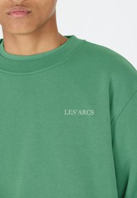 Grön crew neck sweatshirt av mjukt material, med en vit "LES’ARCS" logotyp fram och ribbad krage och ärmar.