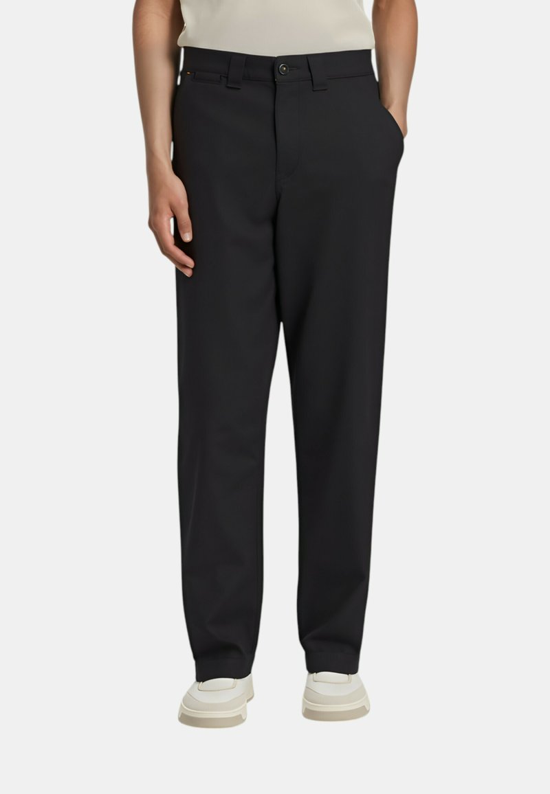 Person wearing black straight-leg trousers with side pockets and white sneakers, standing against plain white background.