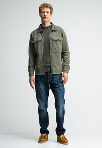 Olijfgroene, gekraagde jas met twee borstzakken, gedragen over een donker gebreid vest. Gecombineerd met blauwe jeans en tan suède schoenen.