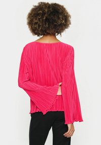 Svetlo roza, pliseirana bluzica s širokimi zvonastimi rokavi in okroglim izrezom. Tekstura je gubana, kar dodaja globino oblikovanju.