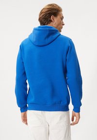 Royalblå hoodie med dragsko, ribbad ärmslut och nederkant. Har en mjuk textur och en avslappnad passform. Ingen synlig branding.