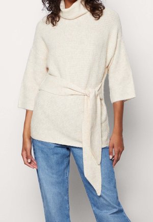 Femme portant un pull en tricot couleur crème avec une ceinture nouée à la taille et des manches trois-quarts, associé à un jean bleu.