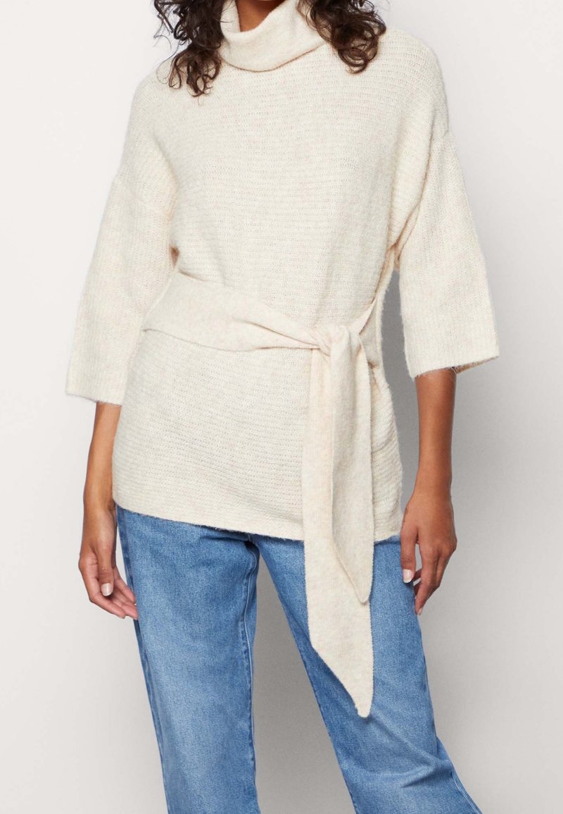 Femme portant un pull en tricot couleur crème avec une ceinture nouée à la taille et des manches trois-quarts, associé à un jean bleu.