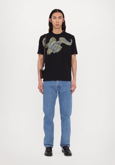 T-shirt noir avec un motif abstrait multicolore, associé à un jean droit bleu clair et des chaussures noires. Accessoirisé avec un sac marron.