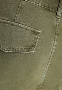 Tela de denim verde oliva, con una vista en primer plano de un bolsillo, costuras visibles y una textura suave. Los acentos incluyen líneas sutiles.