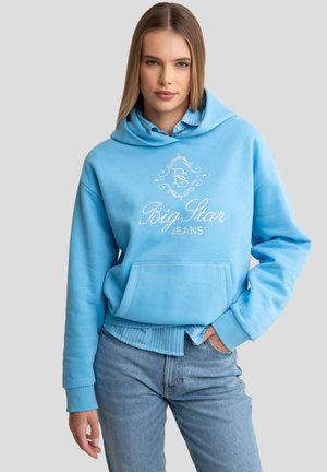 Helleblaue Kapuzenjacke mit Fronttasche, versehen mit einem weißen Logo und dekorativem Text. Kombiniert mit einem hellblau gestreiften Hemd und Jeans.