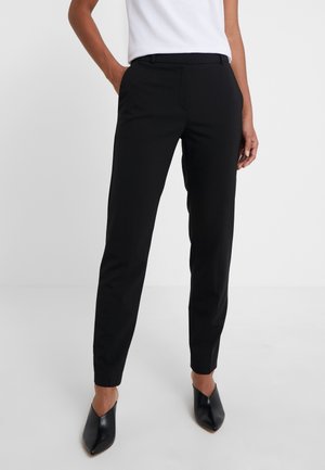 Femme portant un pantalon noir ajusté, un haut blanc sans manches et des chaussures noires à bouts pointus, debout avec une main dans la poche.