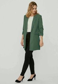 Blazer verde con un design su misura, maniche lunghe e due tasche anteriori, abbinato a una maglietta bianca, pantaloni neri e décolleté nere.