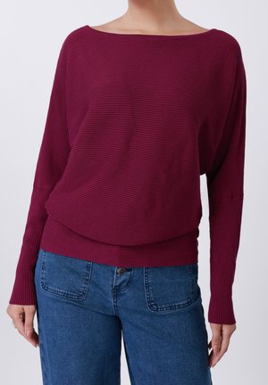Femme portant un haut bordeaux côtelé à manches longues et épaules dénudées, associé à un jean bleu taille haute avec de grandes poches avant.