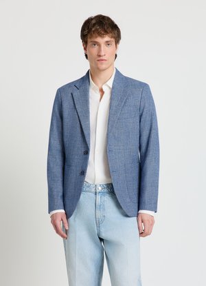 Junger Mann, der einen blau strukturierten Blazer über einem weißen Hemd und hellblauen Jeans trägt, steht vor einem schlichten weißen Hintergrund.