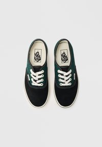 Svarta mockaskor med en grön leopardmönstrad kanvastopp, vita snören och kontrasterande sömmar. Klassisk Vans-logotyp närvarande.