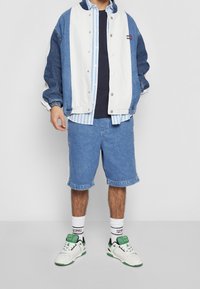 Chaqueta de mezclilla con bloques de color blanco y azul, superpuesta a una camisa de rayas azul, combinada con pantalones cortos de mezclilla y zapatillas blancas con acentos verdes.