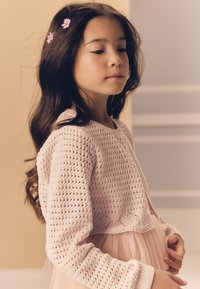 Maglione lavorato a maglia di colore rosa chiaro con un motivo testurizzato, indossato sopra un abito in tulle rosa chiaro. Capelli lunghi e scuri con fermagli a forma di fiore su un lato.