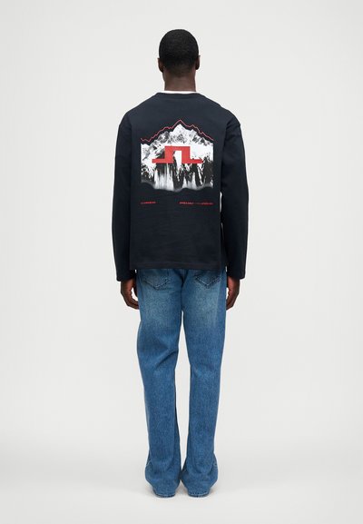 Férfi fekete hosszú ujjú pólóban, hegyi grafikával és piros dizájnnal, kék farmernadrágban áll egy sima fehér háttér előtt.