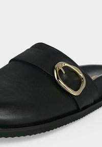 Μαύρο δερμάτινο παπούτσι slip-on με χρυσή αγκράφα ακανόνιστου σχήματος στο πλάι και υφή στην σόλα.