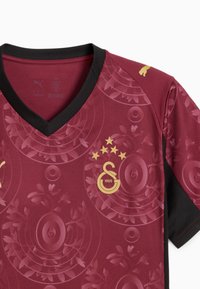 Weinrotes Fußballtrikot mit V-Ausschnitt, schwarzen Akzenten und strukturiertem Muster mit kreisförmigen und blumigen Motiven. Goldene Logos und Verzierungen.