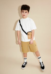 Camiseta blanca de manga corta, pantalones cortos tipo cargo color caqui, zapatillas negras con suelas y cordones blancos, calcetines de rayas blancas y negras, y un pequeño bolso marrón.