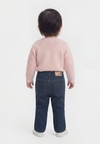 Kleinkind mit kurzem dunklem Haar steht mit dem Rücken zum Betrachter, trägt einen rosa Pullover, dunkelblaue Jeans und weiße Schuhe vor einem einfarbigen Hintergrund.