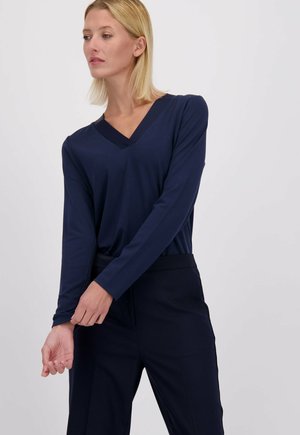 Donkerblauwe lange mouw top met een V-hals, gemaakt van gladde stof. Het heeft een relaxte pasvorm en aangesloten mouwen, gecombineerd met donkere broek.